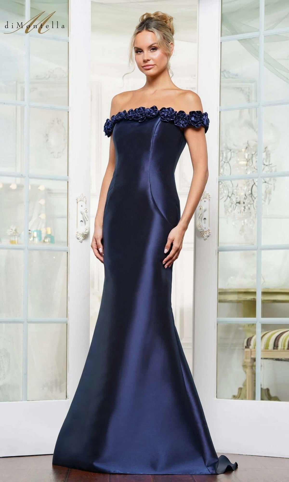 A woman stands in an elegant white room wearing the Long Formal Dress: Rina Di Montella RD3205, a floor-length navy gown with off-the-shoulder design and floral neckline detailing.